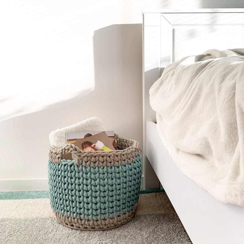 May include: A large, crocheted storage basket with a beige and teal color scheme. The basket is filled with folded blankets and books. The basket is sitting on a light gray rug in front of a white bed.