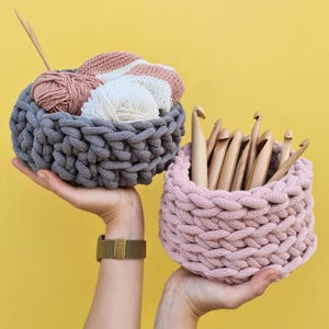 May include: Two crocheted baskets, one gray and one pink, with yarn and crochet hooks inside. The gray basket is filled with yarn balls and a crocheted project. The pink basket is filled with crochet hooks.
