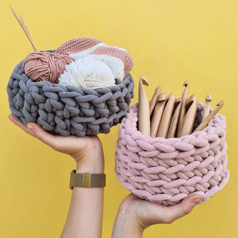May include: Two crocheted baskets, one gray and one pink, with yarn and crochet hooks inside. The gray basket is filled with yarn balls and a crocheted project. The pink basket is filled with crochet hooks.