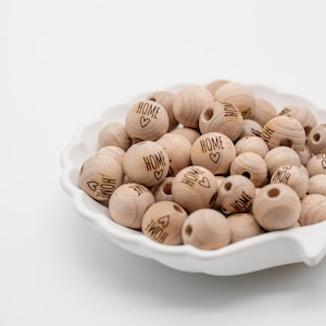 Op de afbeelding: Een witte kom gevuld met houten kralen. Elke kraal is gegraveerd met het woord "HOME" en een klein hartje.