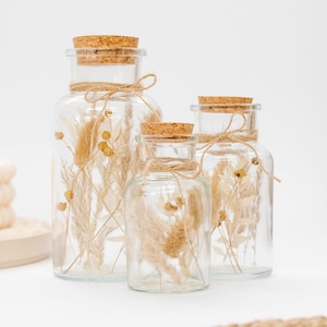 Op de afbeelding: Drie glazen potten met kurken deksels en touw om de hals gebonden. Elke pot bevat gedroogd pampasgras en andere gedroogde bloemen.