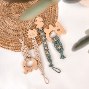 Op de afbeelding: Drie houten bijtringen met macramé-bandjes en houten kralen. De bijtringen zijn gevormd als een leeuw, een koala en een olifant. De bandjes zijn beige en groen. De kralen zijn gegraveerd met de letters "PAUL", "IMRAN" en "LILA".