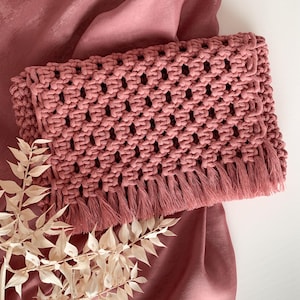 May include: A dusty rose macrame clutch bag with a fringe detail. The bag is made with a woven knot pattern.
