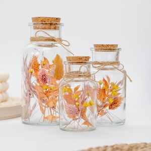 Op de afbeelding: Drie glazen potten met kurken deksels en touw om de hals gebonden. Elke pot bevat gedroogde bloemen in tinten oranje, geel en roze.