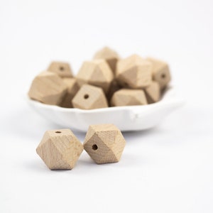 Może przedstawiać: Zbliżenie na dwie geometryczne drewniane koraliki z otworem w środku. Koraliki są ostre i leżą na białym tle. W tle znajdują się inne koraliki, rozmyte, w białej misce.