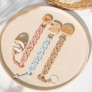 Può includere: Una collezione di clip per ciuccio per bambini in macramè rosa, blu e beige con abbellimenti in legno. Gli accenti in legno includono un cuore, un elefante e un arcobaleno. È presente anche un ciuccio bianco con un cordino rosa.