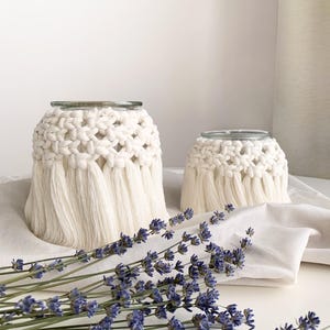 May include: Two white crocheted candle holders with fringe, one larger than the other, on a white surface with lavender flowers.