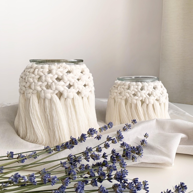 May include: Two white crocheted candle holders with fringe, one larger than the other, on a white surface with lavender flowers.