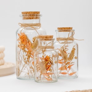 Op de afbeelding: Drie heldere glazen flessen van verschillende formaten, elk met een kurken stop en vastgebonden met touw. De flessen zijn gevuld met gedroogde oranje en beige bloemen en bladeren, wat een rustieke, decoratieve weergave creëert.