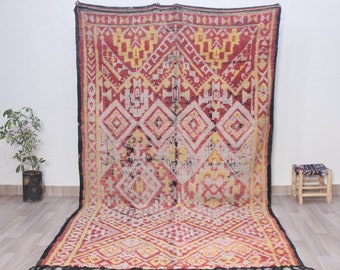 Puede incluir: Una alfombra marroquí vintage con un patrón geométrico en tonos rojos, amarillos y negros. La alfombra tiene un borde de flecos y está tejida con una técnica tradicional.