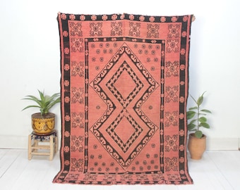 Può includere: Un tappeto vintage rosa e nero con un motivo geometrico. Il tappeto ha una forma di diamante al centro con un bordo nero e un motivo ripetitivo di stelle e fiori.