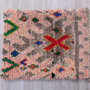 Pu&ograve; includere: Un tappeto marocchino rosa e grigio con un motivo geometrico e accenti colorati. Il tappeto ha un bordo a frange ed &egrave; fatto di lana.