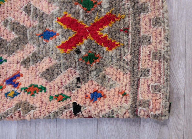 Pu&ograve; includere: Un tappeto rosa e grigio con dettagli ricamati colorati. Il tappeto ha un motivo geometrico e un bordo a frange.