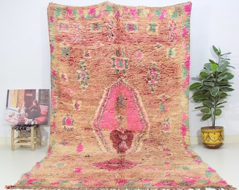 May include: A large, rectangular rug with a textured, woven design. The rug features a central pink geometric shape, surrounded by a beige background with pink and green accents. The rug is displayed against a white background.