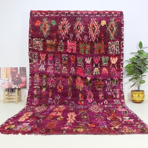May include: A vibrant, hand-woven Moroccan rug with a deep burgundy background and intricate floral patterns in shades of pink, green, and blue. The rug has a fringed edge and a textured, hand-knotted surface.