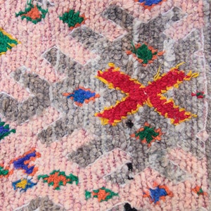 Pu&ograve; includere: Un tappeto marocchino rosa, grigio, verde, blu, rosso e giallo con un motivo geometrico. Il tappeto &egrave; fatto di lana e ha una texture morbida e soffice.