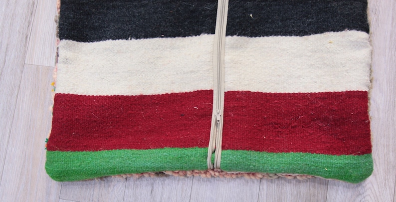 Pu&ograve; includere: Un cuscino a righe colorate con una chiusura a zip. Il cuscino &egrave; realizzato in tessuto tessuto con strisce nere, bianche, rosse e verdi.