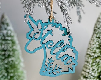 May include: Teal blue wooden ornament shaped like a unicorn with the name "Stella" cut out in the design. The ornament is hanging from a silver and gold string.