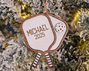 May include: A wooden pickleball paddle ornament with a natural wood border and a white center. The paddle features the engraved text MICHAEL and 2025, along with a small pickleball graphic. It hangs from a thin brown twine loop for display.