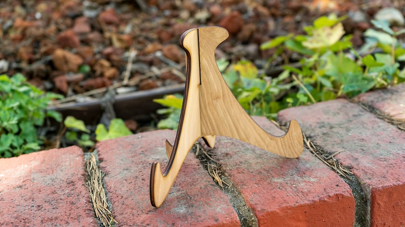May include: A wooden display stand with a natural wood finish. The stand has a triangular shape with three legs and a curved top.
