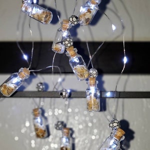 May include: A string of white fairy lights with small glass bottles filled with seashells hanging from them. Each bottle has a black and white patterned bead attached to it.