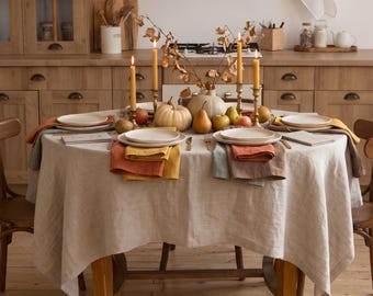 May include: A round dining table covered with a beige linen tablecloth, set with white plates, layered orange and green napkins, and gold candlesticks with lit yellow candles. A centerpiece of small pumpkins, pears, and dried branches sits in the middle.