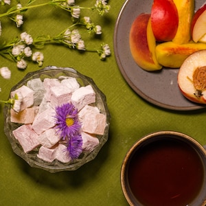 May include: Overhead shot of a table setting with a green tablecloth. A glass bowl holds pink Turkish delight, garnished with purple flowers. Sliced peaches and a cup of tea are also present, along with white flowers.