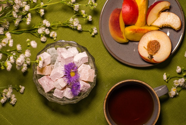 May include: Overhead shot of a table setting with a green tablecloth. A glass bowl holds pink Turkish delight, garnished with purple flowers. Sliced peaches and a cup of tea are also present, along with white flowers.