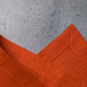 May include: A vibrant burnt orange fabric napkin featuring a mitered corner detail with visible stitching along the hem. The textile has a distinct woven texture and is folded to showcase the clean, sharp corner construction and finished edges.
