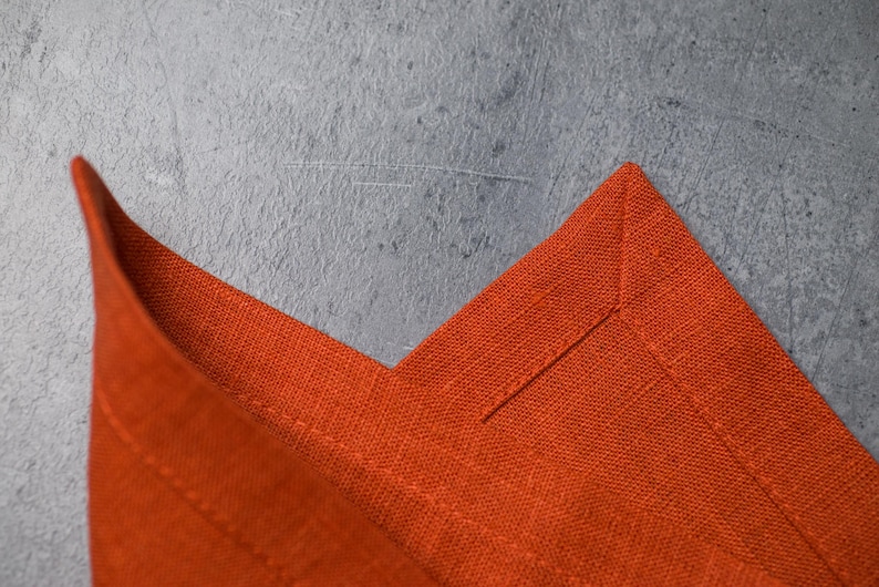 May include: A vibrant burnt orange fabric napkin featuring a mitered corner detail with visible stitching along the hem. The textile has a distinct woven texture and is folded to showcase the clean, sharp corner construction and finished edges.