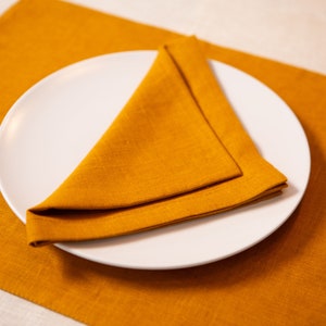 May include: A white ceramic plate with a folded orange napkin on top, set on an orange linen placemat. The napkin is folded into a triangle shape. The image is well-lit, highlighting the textures and colors.