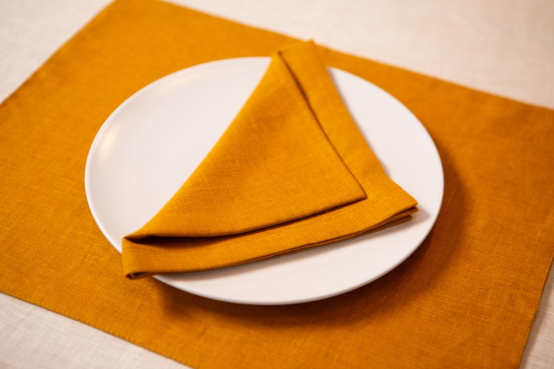 May include: A white ceramic plate with a folded orange napkin on top, set on an orange linen placemat. The napkin is folded into a triangle shape. The image is well-lit, highlighting the textures and colors.