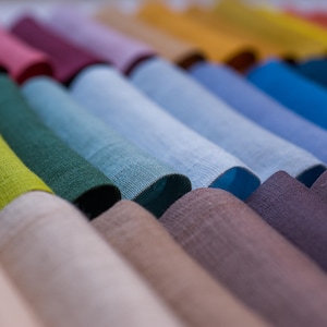 May include: A collection of folded fabric in various colors, including shades of green, blue, purple, and yellow. The textiles appear to be linen or a similar woven material, with a slightly textured surface. The image showcases a range of hues, from light to dark.