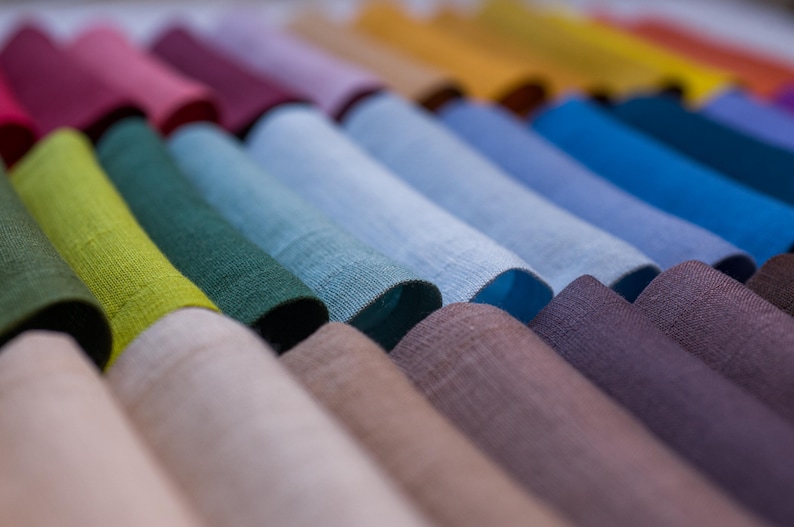 May include: A collection of folded fabric in various colors, including shades of green, blue, purple, and yellow. The textiles appear to be linen or a similar woven material, with a slightly textured surface. The image showcases a range of hues, from light to dark.