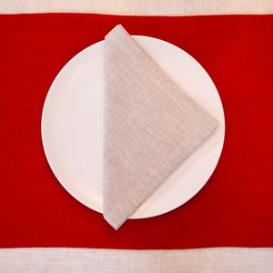 May include: A white plate with a folded linen napkin on a red placemat. The placemat is rectangular and the napkin is folded into a triangle. The background is a light beige tablecloth.