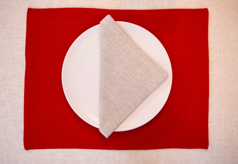 May include: A white plate with a folded linen napkin on a red placemat. The placemat is rectangular and the napkin is folded into a triangle. The background is a light beige tablecloth.