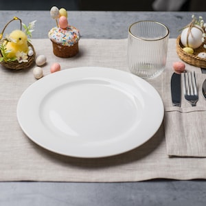 May include: A festive Easter table setting with a white plate, silverware, and a clear glass with a gold rim. Decorations include a chick in a basket, a cupcake with egg-shaped candies, and decorative eggs in a basket. The setting is on a beige linen placemat.