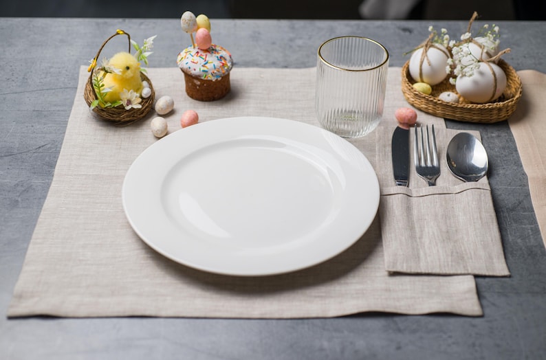May include: A festive Easter table setting with a white plate, silverware, and a clear glass with a gold rim. Decorations include a chick in a basket, a cupcake with egg-shaped candies, and decorative eggs in a basket. The setting is on a beige linen placemat.