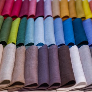 May include: A collection of folded linen napkins in a spectrum of colors, including red, pink, green, blue, and purple. The napkins are arranged in rows, showcasing a variety of hues and textures. The image highlights the diverse color palette.