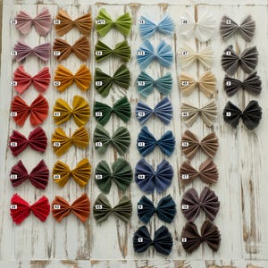 May include: A collection of pleated fabric bows arranged in a grid, each featuring a small white square label with a black number. The bows are displayed in a wide spectrum of colors including pink, orange, green, blue, white, gray, and brown.