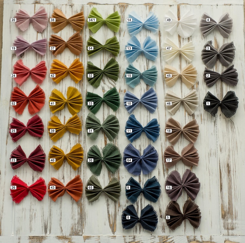 May include: A collection of pleated fabric bows arranged in a grid, each featuring a small white square label with a black number. The bows are displayed in a wide spectrum of colors including pink, orange, green, blue, white, gray, and brown.