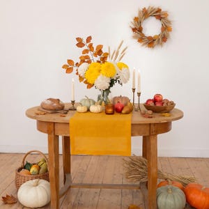 Pode incluir: Uma mesa de jantar de madeira com um caminho de mesa amarelo mostarda, um vaso de vidro com crisântemos amarelos e brancos, espigas de trigo e folhas de outono. A mesa contém velas, pequenas abóboras, peras, maçãs e um pão.
