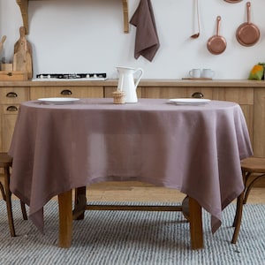 Op de afbeelding: Een mauve linnen tafelkleed gedrapeerd over een ronde houten tafel met twee houten stoelen. De tafel is gedekt met twee witte borden en een witte kan.