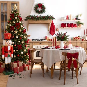 Könnte beinhalten: Eine große Nussknacker-Figur steht neben einem geschmückten Weihnachtsbaum mit goldenen und roten Kugeln, roten Schleifen und einem Stern. Ein Esstisch mit beiger Tischdecke, roten Schleifen an den Stühlen und festlicher Dekoration steht in einer Küche.