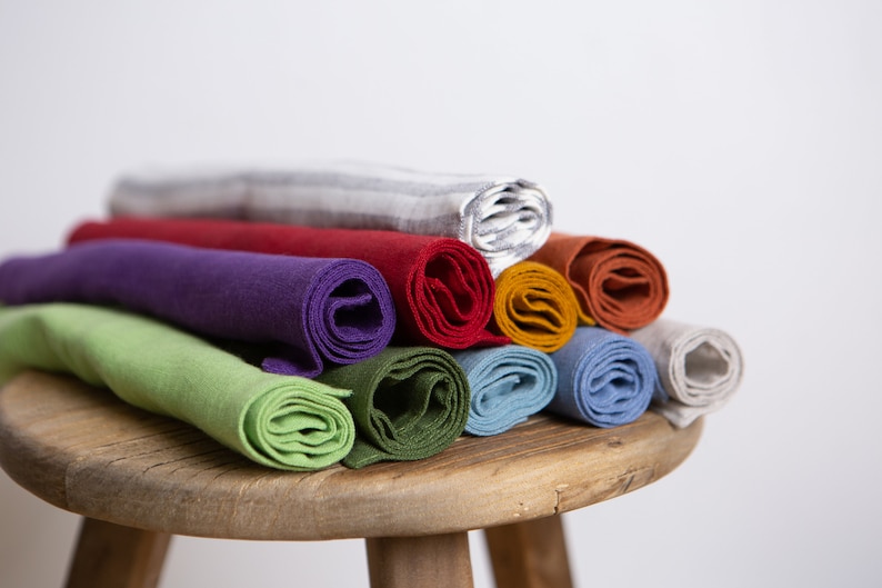 May include: A stack of rolled fabric in various colors, including green, purple, red, and blue. The rolls are arranged on a small, wooden stool. The fabric appears to be soft and possibly linen or cotton.