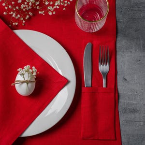 May include: A red linen placemat with a white plate, silverware, and a glass with a gold rim. A white egg with flowers tied with twine sits on the napkin. White flowers are in the upper left corner.