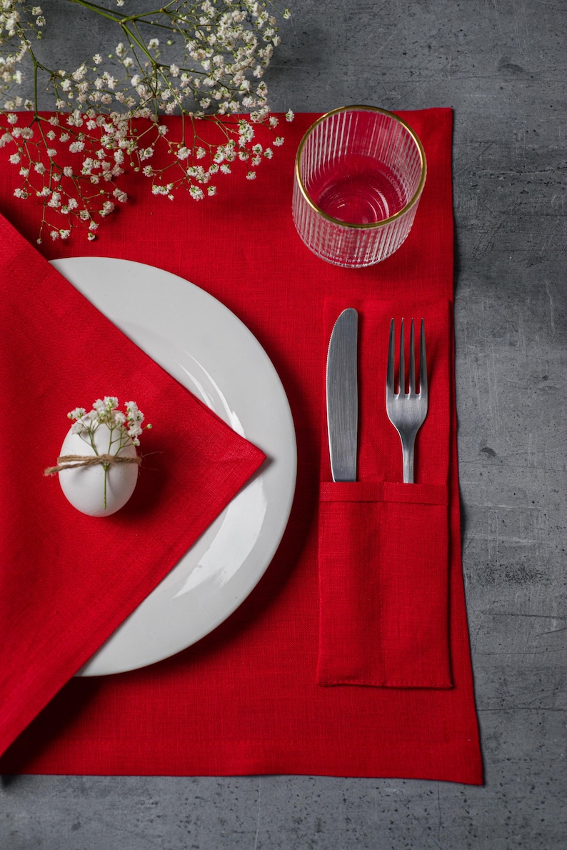 May include: A red linen placemat with a white plate, silverware, and a glass with a gold rim. A white egg with flowers tied with twine sits on the napkin. White flowers are in the upper left corner.