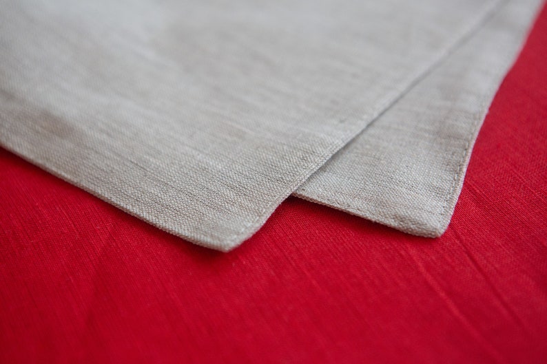 May include: A close-up of two folded linen napkins. The top napkin is a light beige color, and the bottom napkin is a vibrant red. The fabric has a textured appearance, suggesting a natural fiber like linen or cotton. The napkins are likely for dining.
