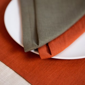 May include: A white ceramic plate with two folded linen napkins. One napkin is olive green, and the other is a burnt orange color. The napkins are placed on a matching burnt orange linen tablecloth.