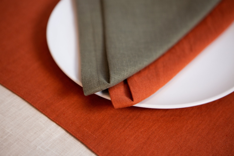 May include: A white ceramic plate with two folded linen napkins. One napkin is olive green, and the other is a burnt orange color. The napkins are placed on a matching burnt orange linen tablecloth.